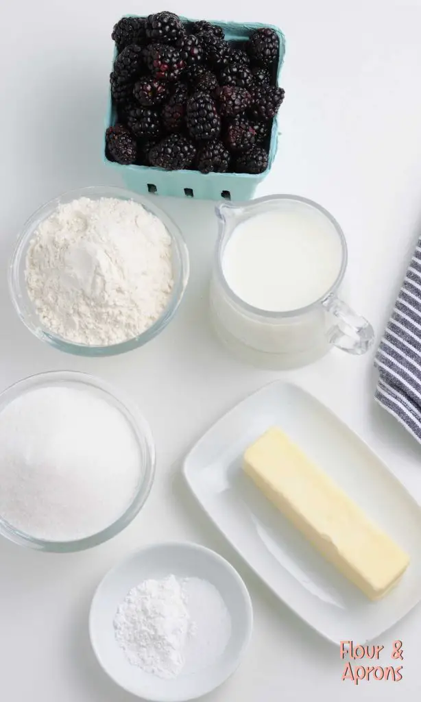 Blackberry cobbler ingredients, from top clockwise around: pint of blackberries, measuring cup of milk, butter, salt & baking powder in small bowl, bowl of sugar, bowl of flour.