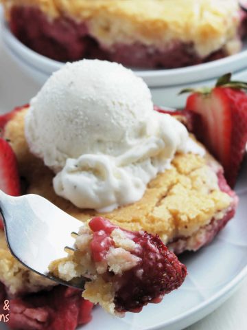 fork with a bite of strawberry cobbler on it, background has a plate with strawberry cobbler and ice cream.