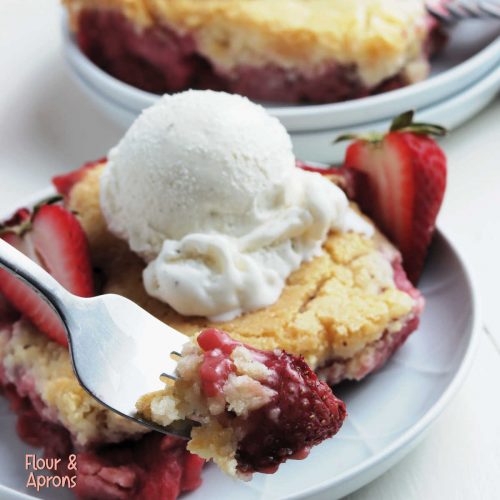fork with a bite of strawberry cobbler on it, background has a plate with strawberry cobbler and ice cream.