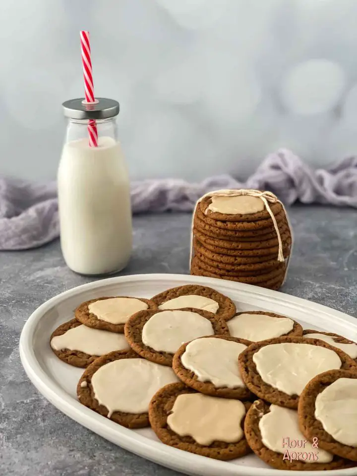 Frosted molasses cookies on a plate with a pile of cookies and a glass of milk in the background.