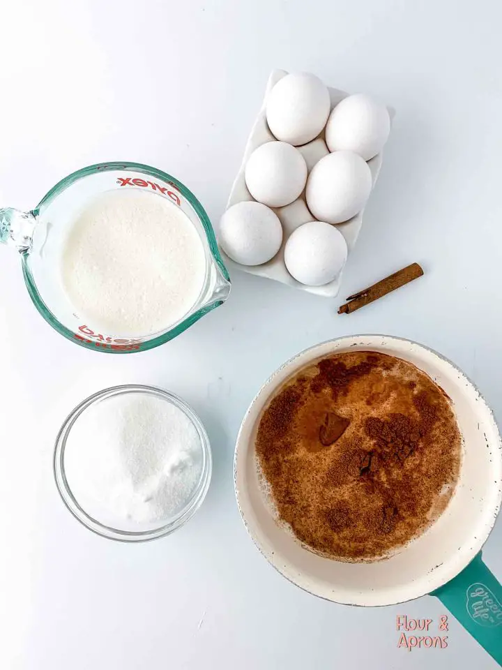Pan with milk, nutmeg, and vanilla extract, small bowl of sugar, measuring dish of milk and egg surrounding it.