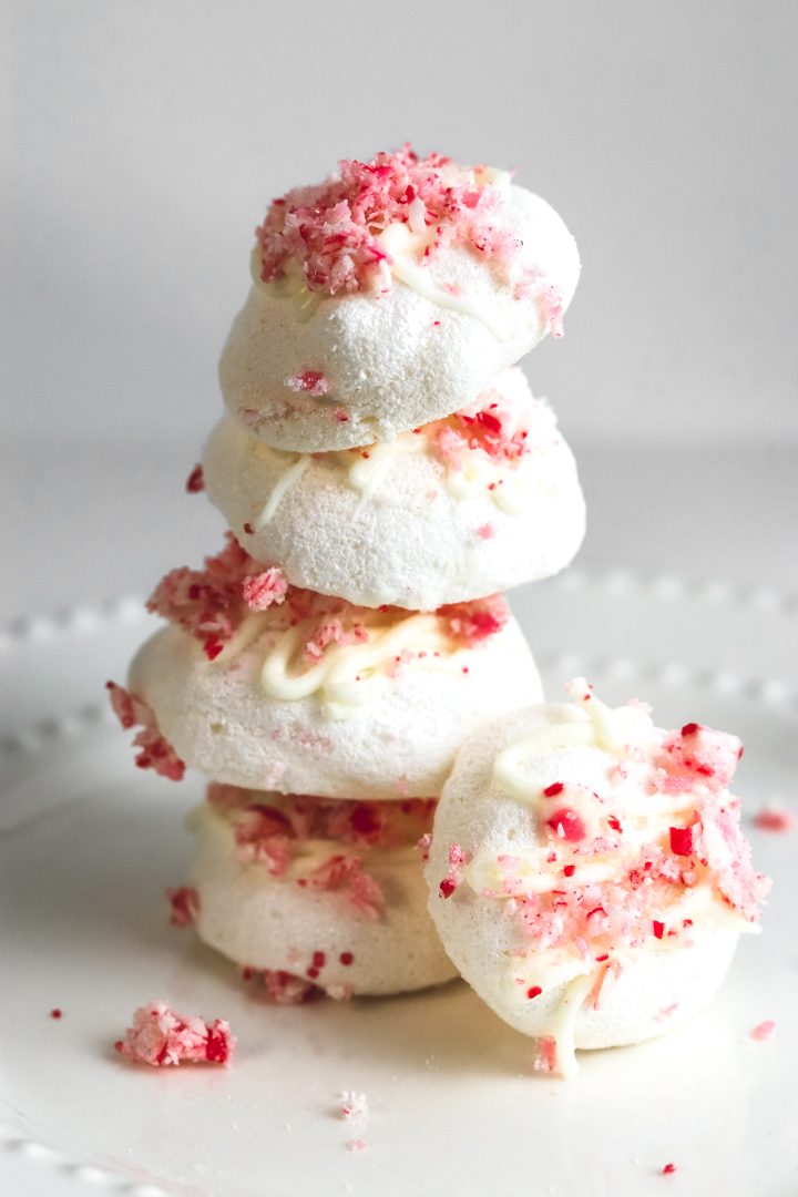 Meringue cookies with drizzled white chocolate and crushed candy canes on top stacked on a plate.