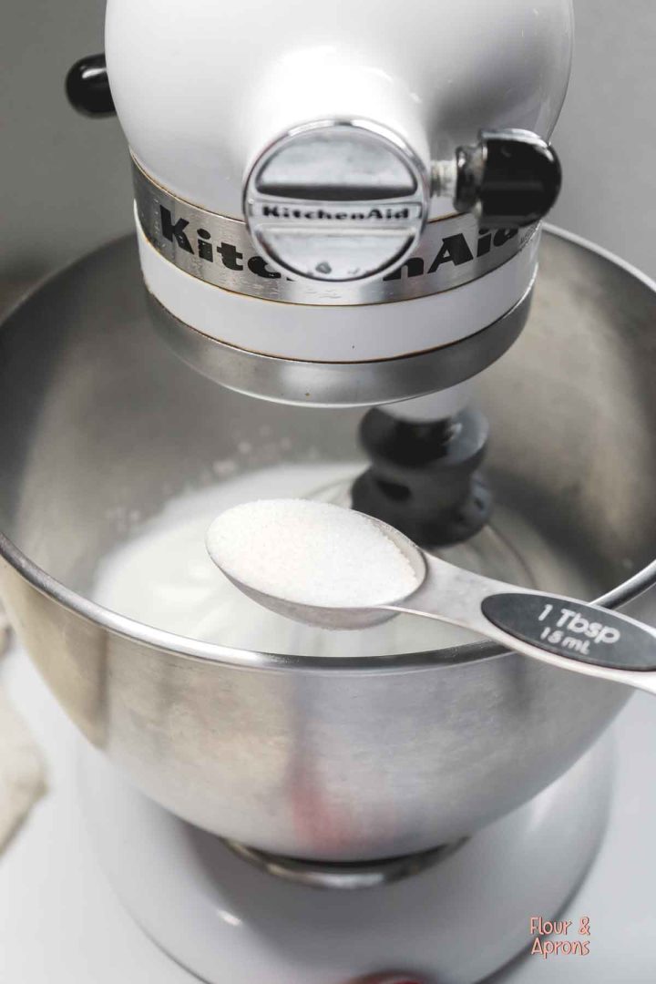 Adding sugar into the stand mixer bowl a Tablespoon at a time.